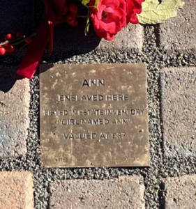 Memorial plaque for Ann, commemorating her history of enslavement, with flowers placed on brick pavement.