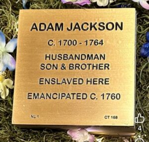 Bronze plaque honoring Adam Jackson, depicting life dates, roles, enslavement, and emancipation, surrounded by flowers.