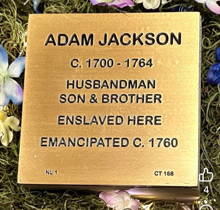 Bronze plaque honoring Adam Jackson, depicting life dates, roles, enslavement, and emancipation, surrounded by flowers.