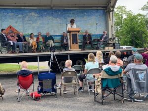 Community gathering with speaker on stage, audience seated outdoors, large painting backdrop.