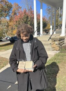 Woman holding plaques outdoors with autumn trees in the background, smiling brightly.