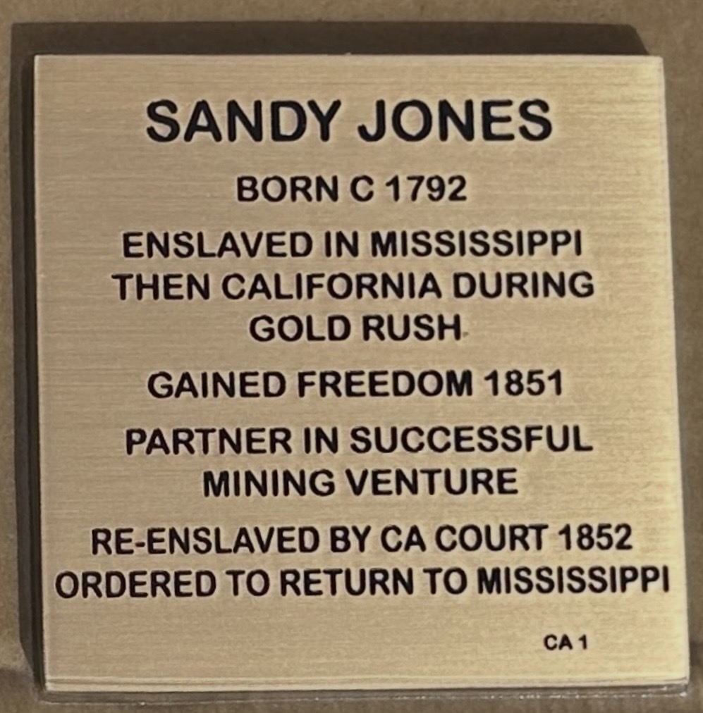 Plaque detailing Sandy Jones' life: born 1792, enslaved in Mississippi, freedom in 1851, mining partner, re-enslaved in 1852.